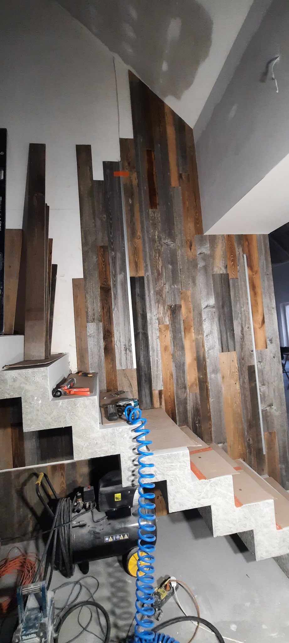 Montaż drewnianej okładziny ściennej na poddaszu, widoczne narzędzia i kompresor, surowe ściany i schody w trakcie wykończenia.