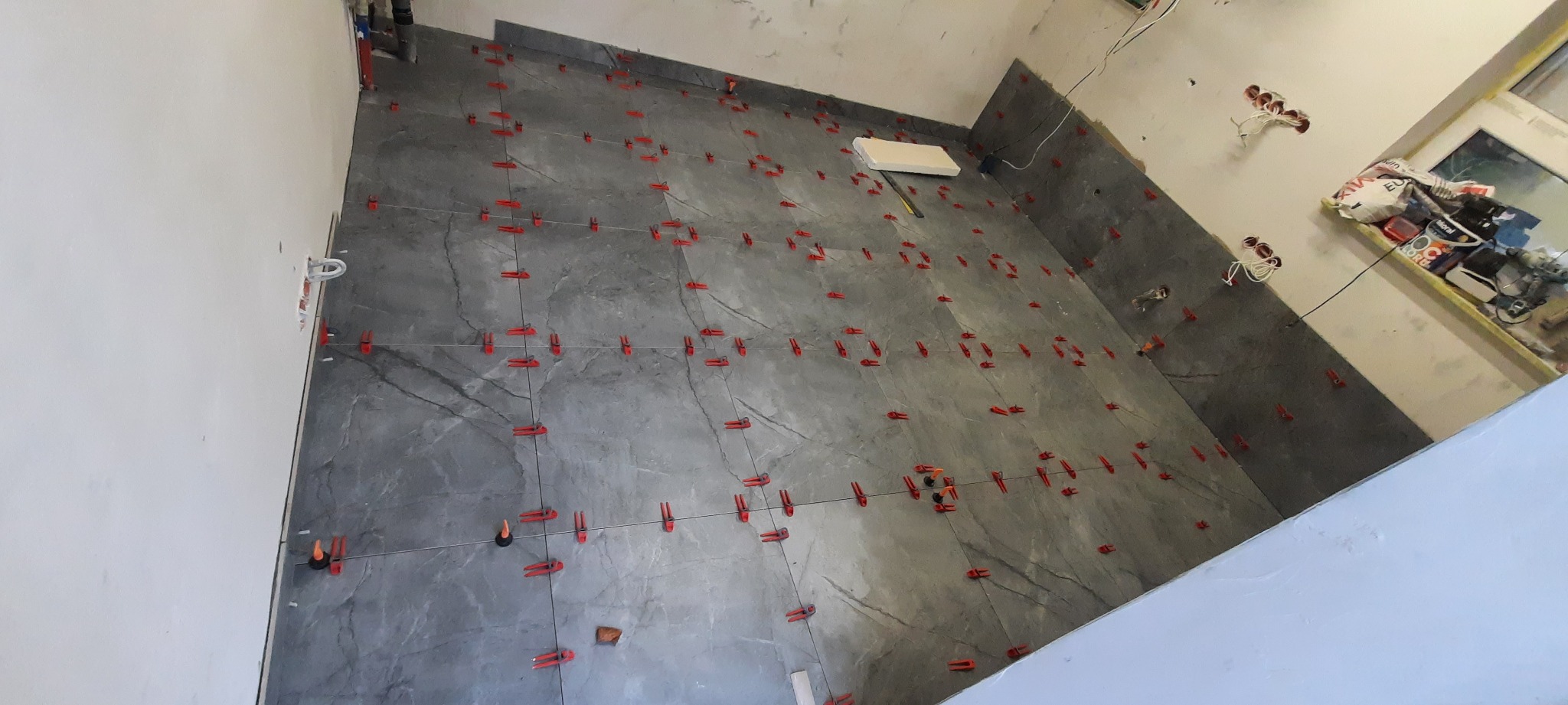 Szare płytki podłogowe z imitacją kamienia, ułożone z użyciem czerwonych klipsów poziomujących, widoczne przewody elektryczne i narzędzia w tle.