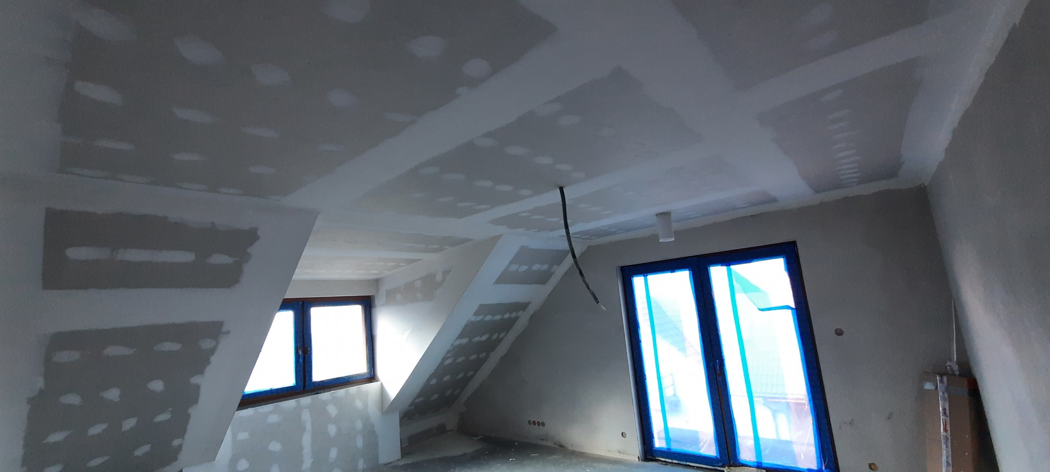 Poddasze w trakcie wykańczania, widoczne płyty kartonowo-gipsowe na ścianach i suficie, zaszpachlowane łączenia, okna zabezpieczone folią malarską, surowy kabel zwisa z sufitu.