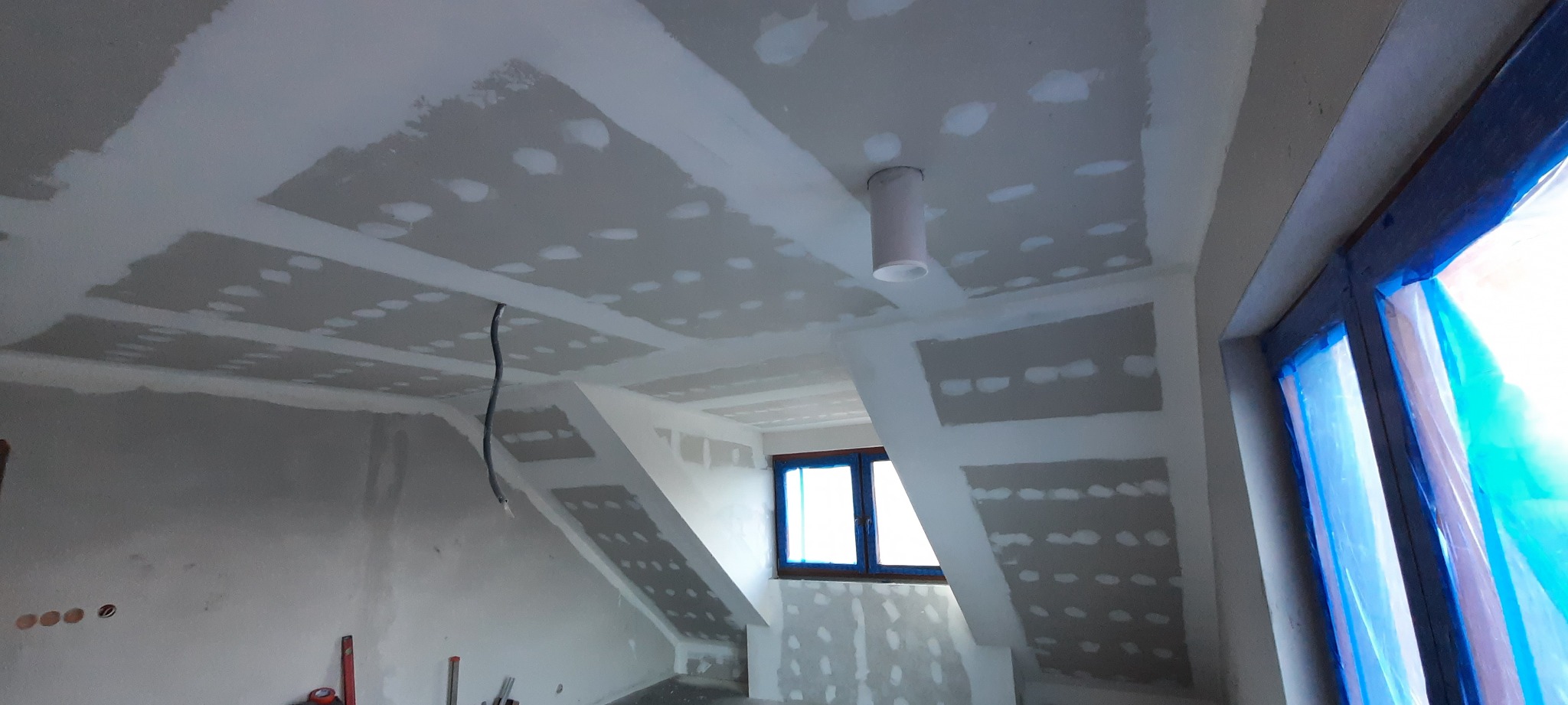 Wykończenie poddasza płytami gipsowo-kartonowymi, widoczne szpachlowane łączenia, sufit z zamontowanym oświetleniem punktowym, okno zabezpieczone folią malarską.