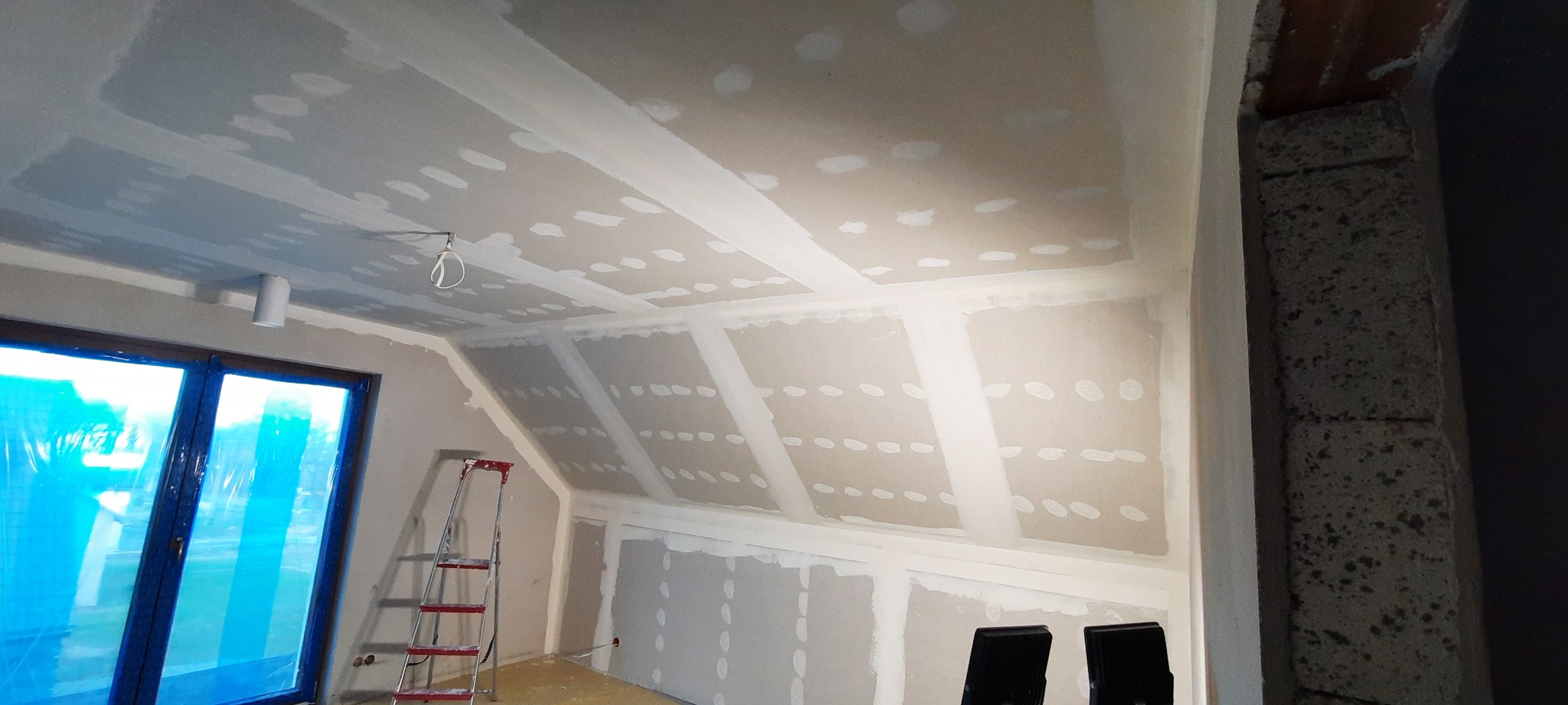 Poddasze w trakcie wykańczania płytami kartonowo-gipsowymi, widoczne łączenia szpachlowane, drabina oparta o ścianę, okno zabezpieczone folią malarską.