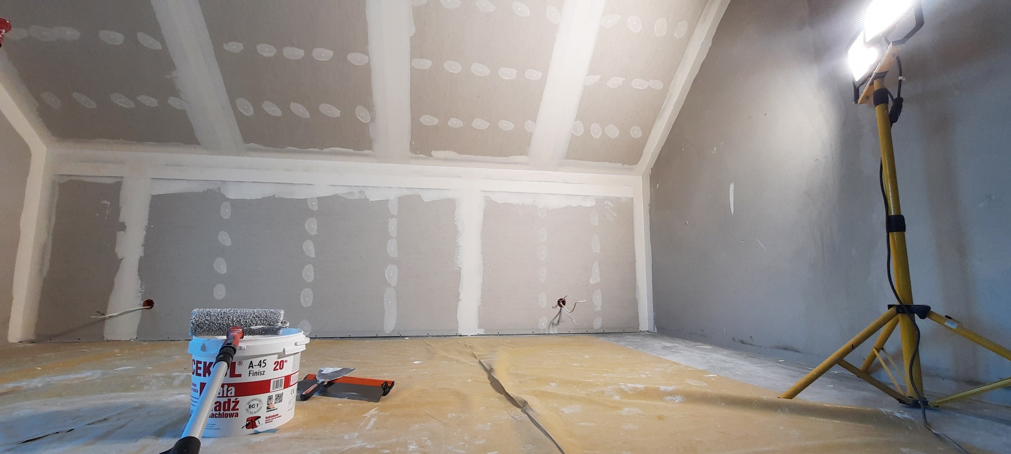 Pokój na poddaszu w trakcie wykańczania: widoczne płyty kartonowo-gipsowe na ścianach i suficie, szpachlowane łączenia, wiadro z farbą i wałek malarski na podłodze zabezpieczonej folią, lampa...