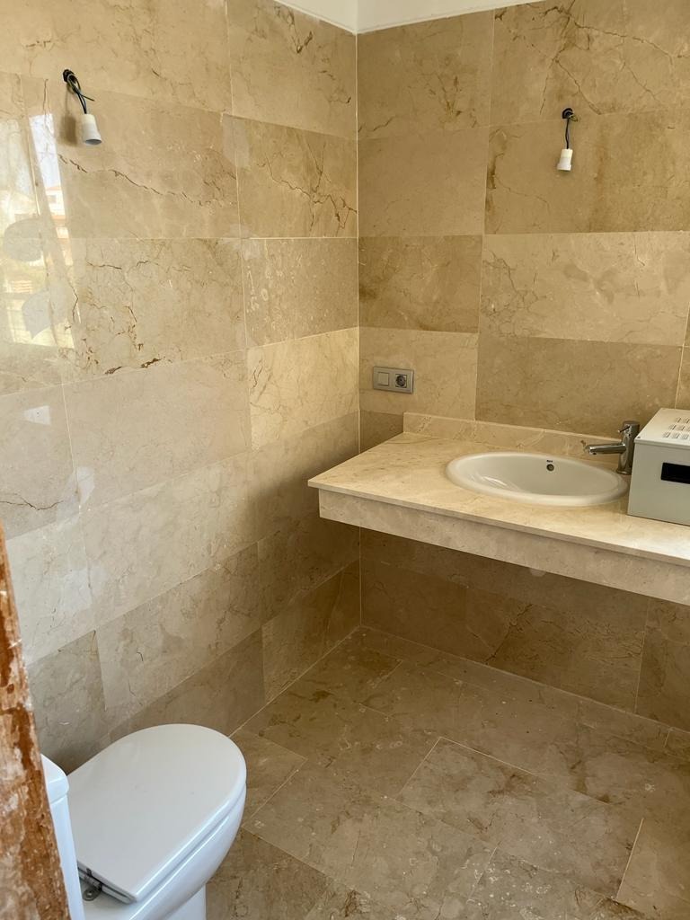 Beżowa łazienka wykończona marmurem, z umywalką wpuszczoną w blat, toaleta w rogu i niedokończone lampy sufitowe.