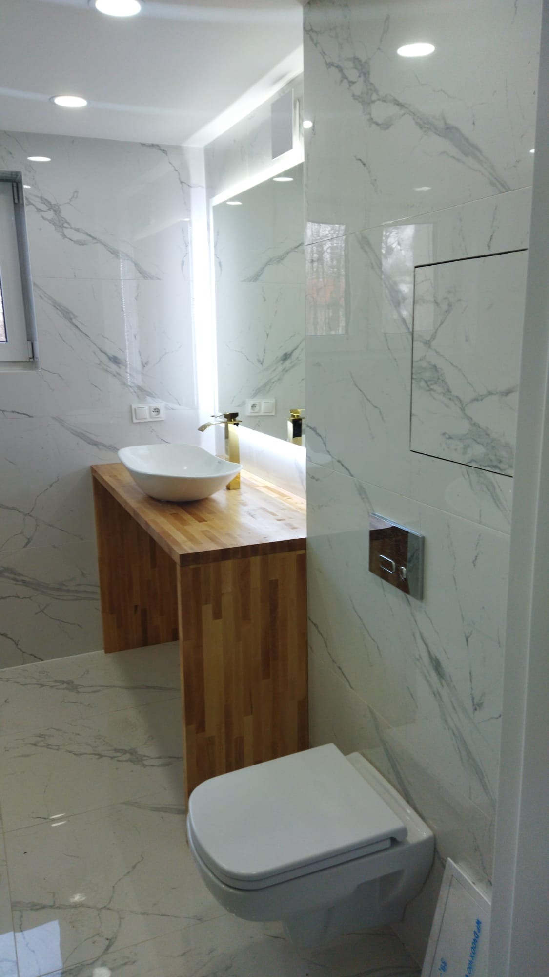Nowoczesna łazienka z białymi marmurkowymi płytkami, drewnianym blatem pod umywalkę nablatową i złotą armaturą, wisząca toaleta.