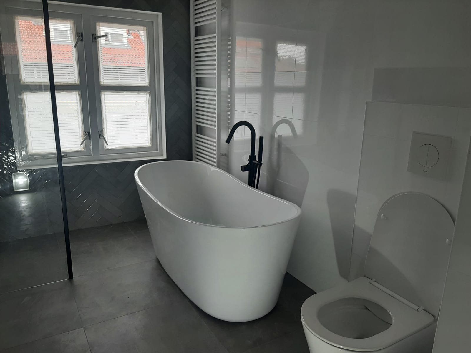 Nowoczesna łazienka z wolnostojącą białą wanną, czarną armaturą, szarą glazurą ułożoną w jodełkę i grzejnikiem drabinkowym.