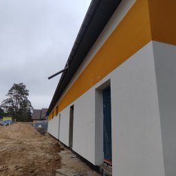 Usługi elektryczne EASY LIGHT Krystian Bałtrukanis - Świeżo otynkowana elewacja budynku z poziomym, żółtym pasem. Okna zabezpieczone folią. Na pierwszym planie hałda piachu, tło stanowią drzewa i szare niebo.