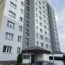 Wysoki budynek mieszkalny po renowacji elewacji, z widocznymi dwoma odcieniami szarości. Na pierwszym planie zaparkowane samochody i wejście do budynku.