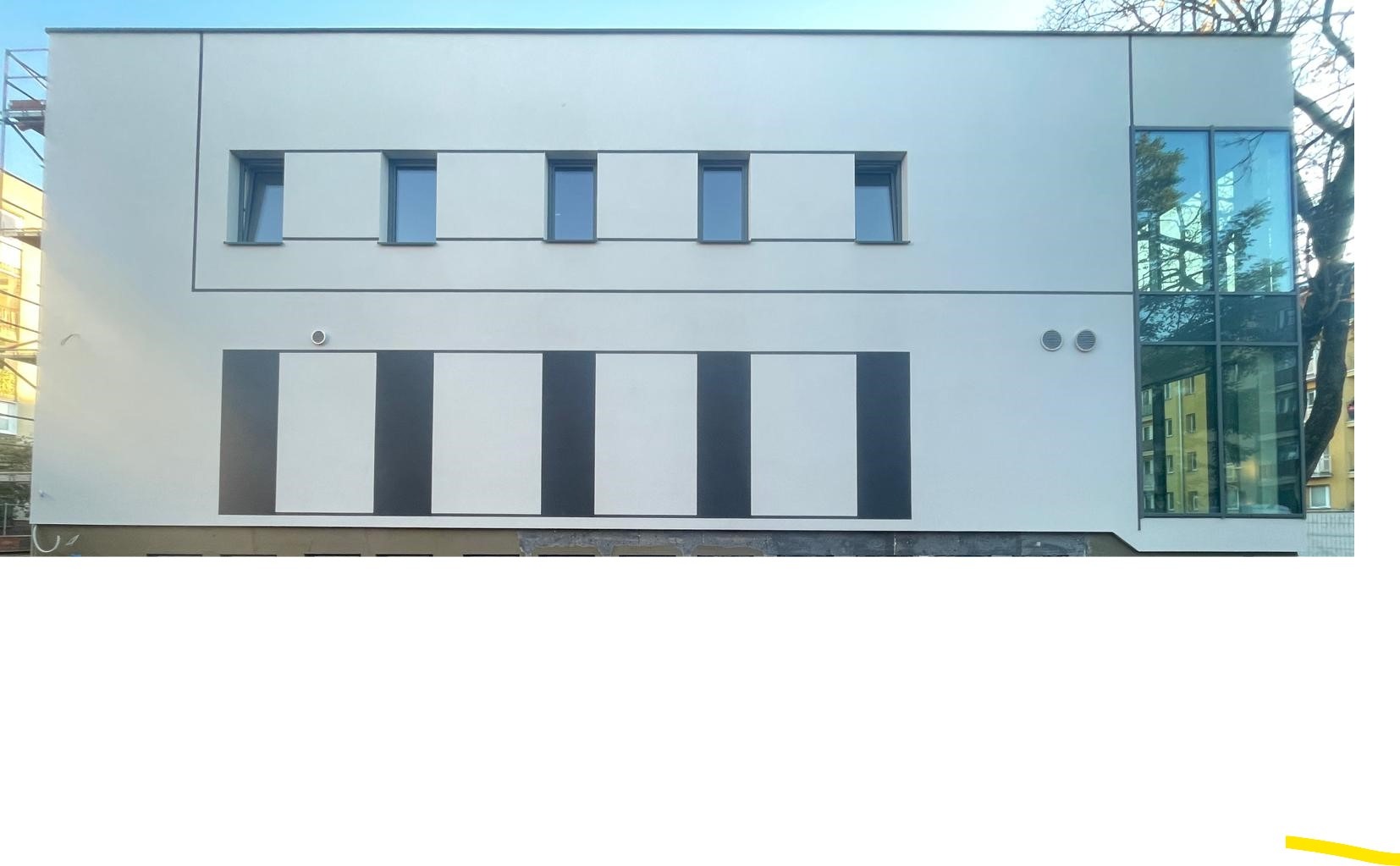 Nowoczesna elewacja budynku usługowego z białymi ścianami, rzędem pięciu okien na górze i pionowymi czarno-białymi pasami na dole, z przeszkloną klatką schodową po prawej stronie i drzewem w tle.