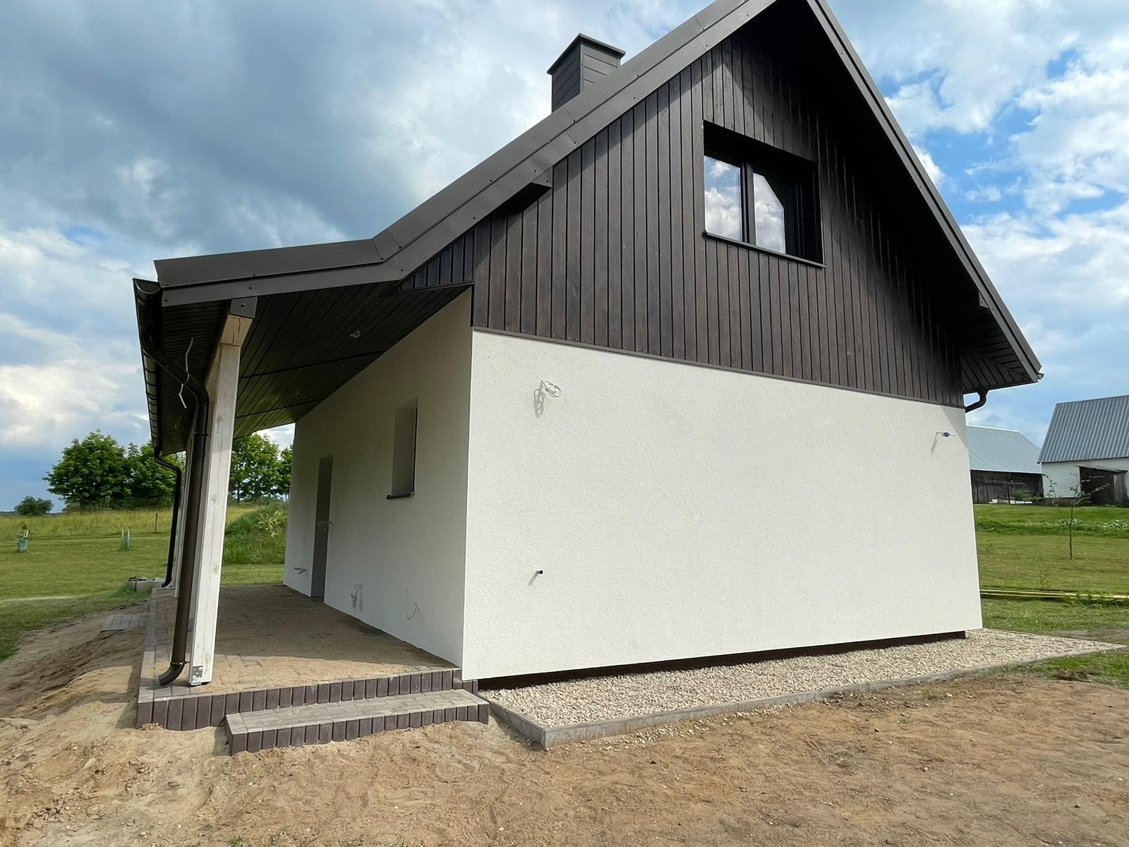 Nowoczesny dom z białą elewacją i ciemnobrązową drewnianą górną częścią oraz podbitką dachu, widoczne niezagospodarowane otoczenie i przygotowane schody wejściowe.