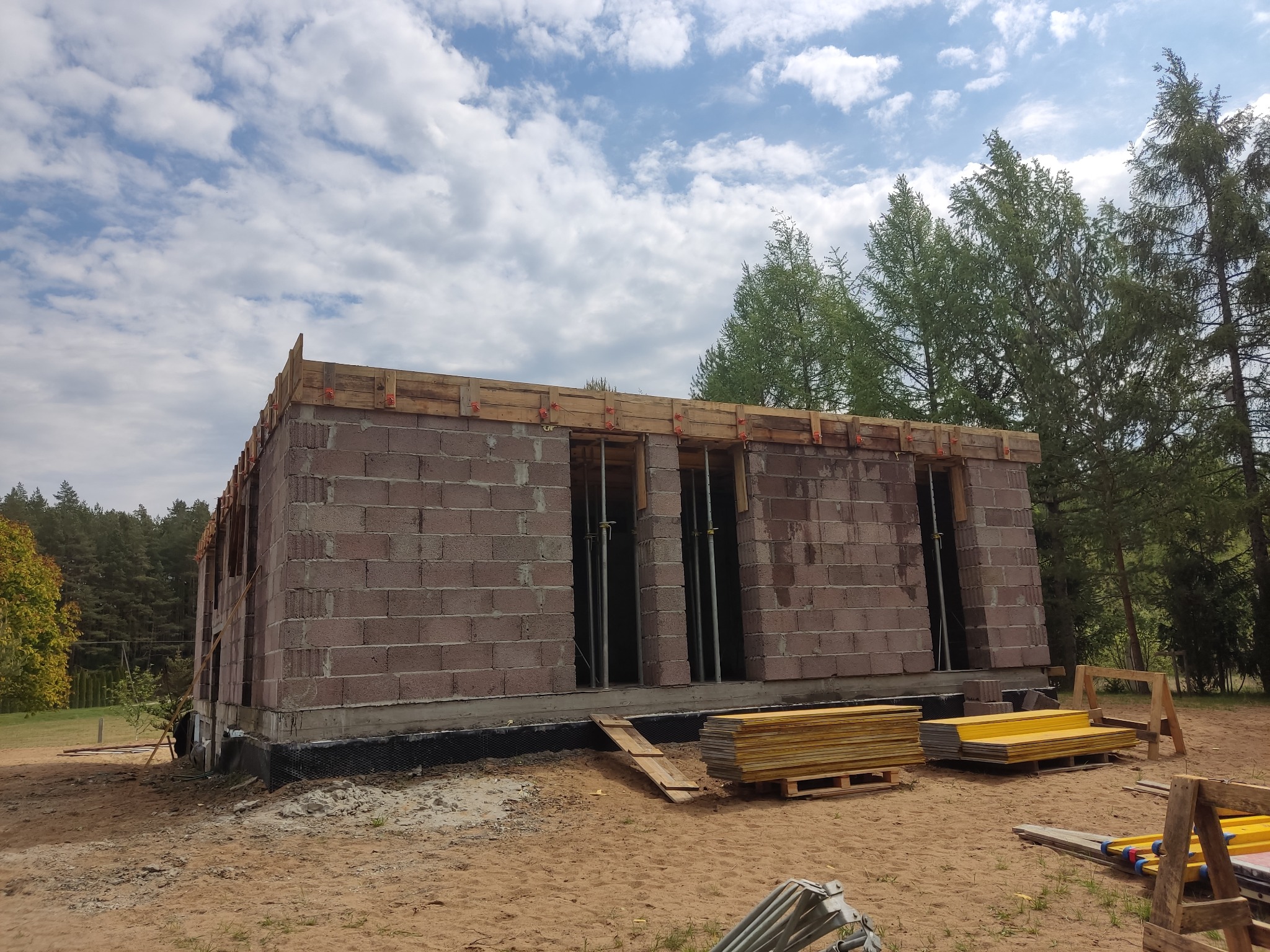 Wznoszenie ścian parterowego domu z bloczków, widoczne drewniane szalunki na górze muru, elementy rusztowania i deski na ziemi. Budowa w otoczeniu drzew i błękitnego nieba.