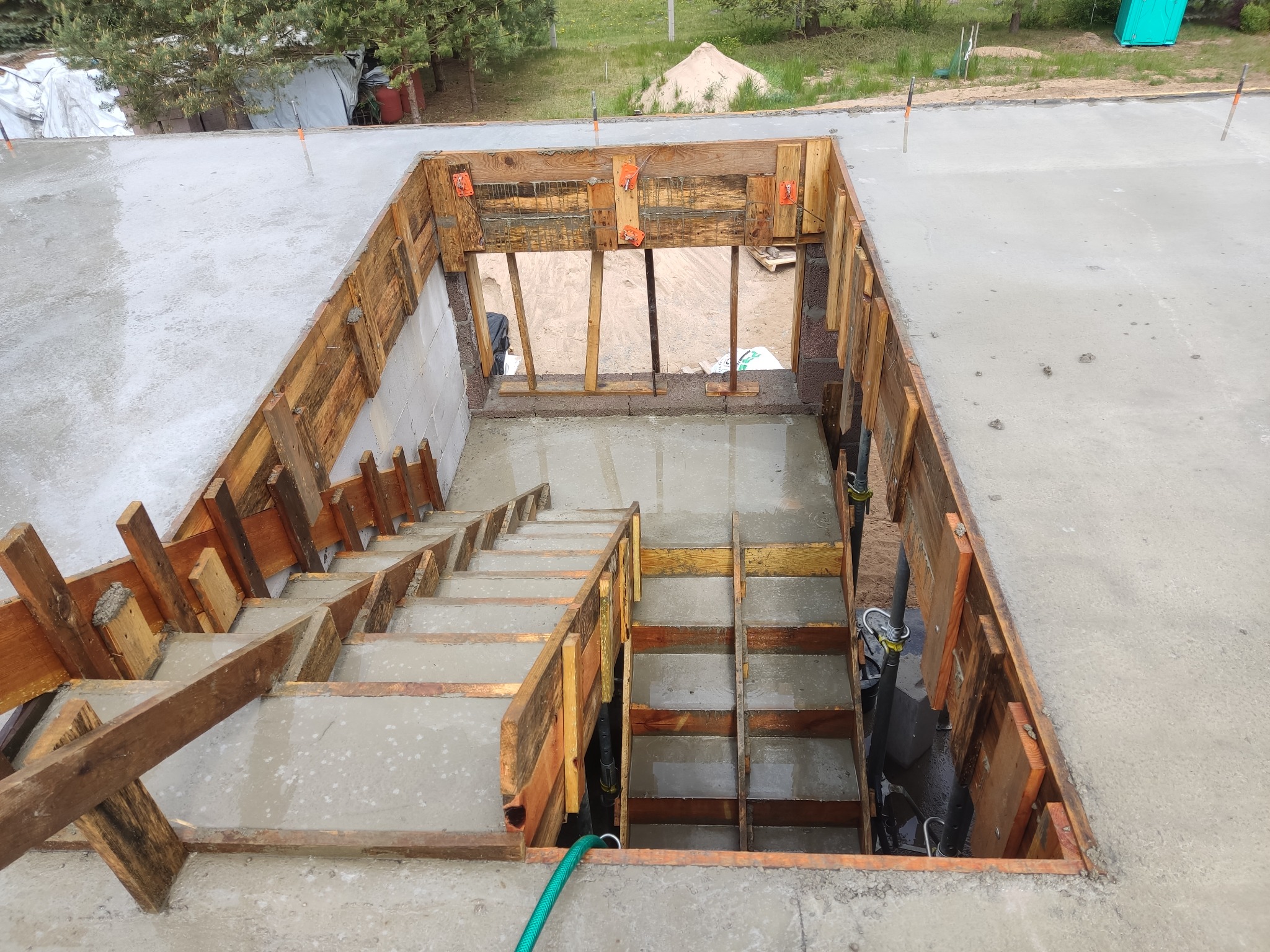 Betonowe schody w trakcie budowy z drewnianym szalunkiem, widok z góry. Świeży beton na stopniach i platformie. W tle widoczny teren budowy.