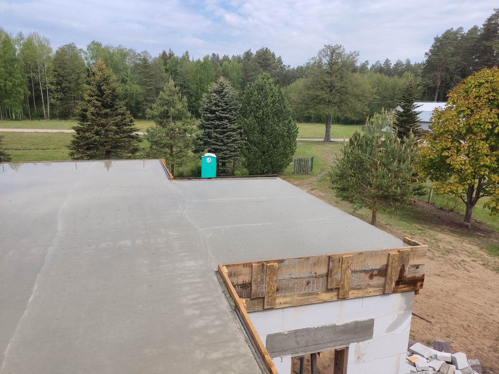 Wylany strop budynku w trakcie budowy, widoczne szalunki i bloczki. W tle las i przenośna toaleta.