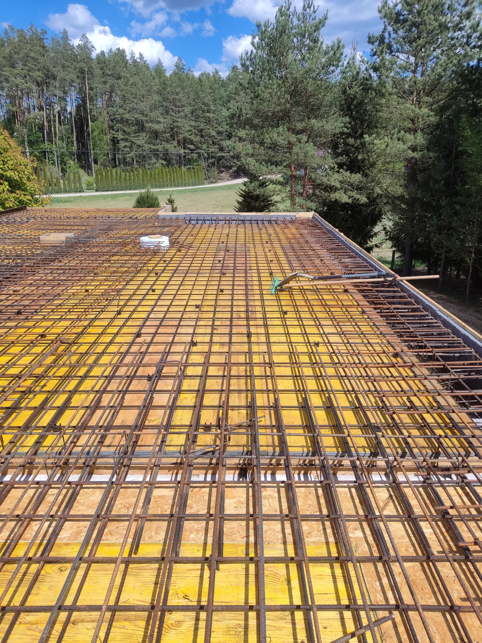 Zbrojenie stropu na żółtym szalunku, widok z góry na siatkę z prętów, narzędzia i otaczającą zieleń drzew. Etap budowy domu.