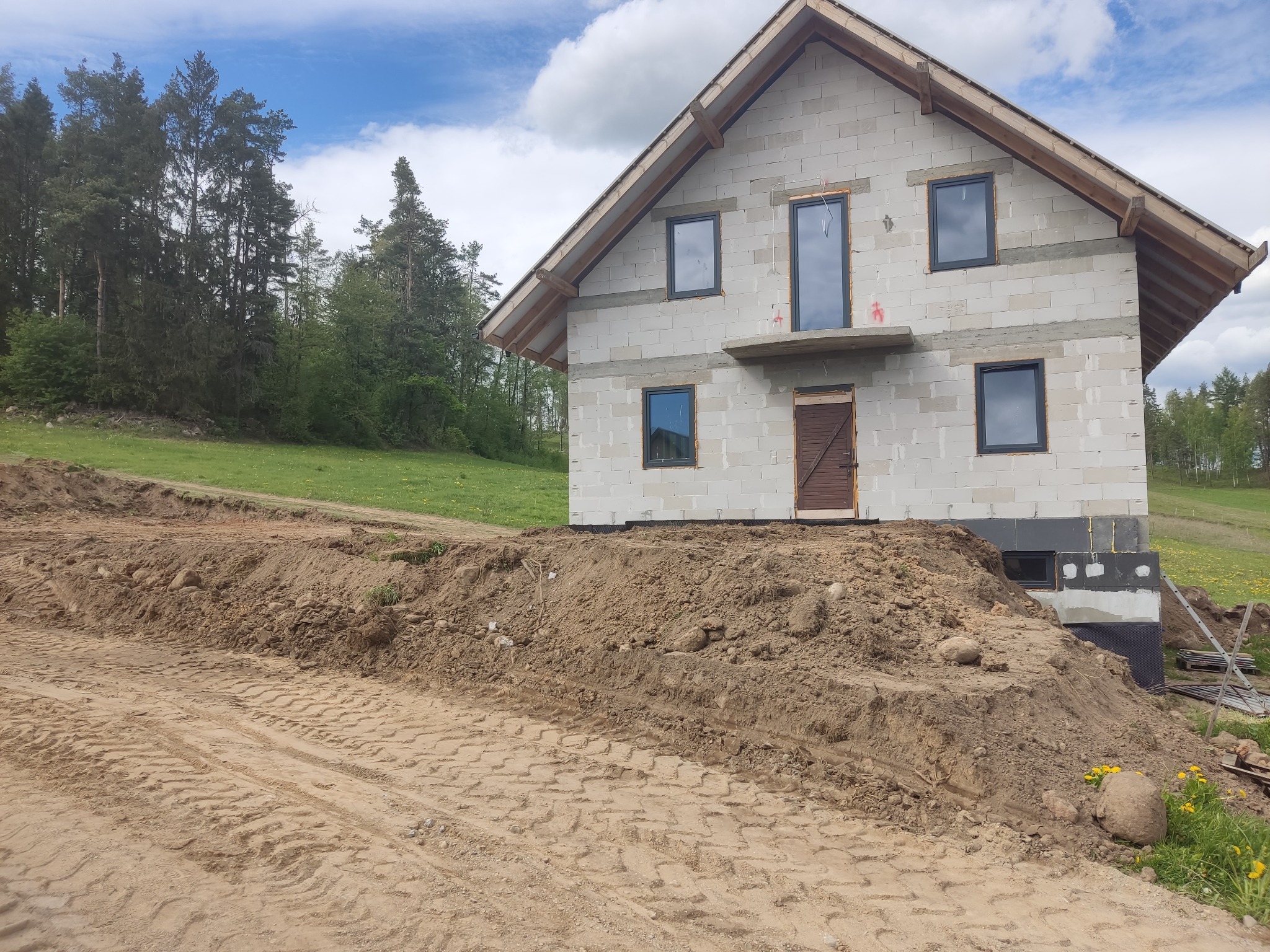 Surowy dom z białego bloczka na wzgórzu z widocznymi śladami opon, okna w ciemnych ramach, drewniane elementy dachu, zielone drzewa w tle, błękitne niebo z chmurami.