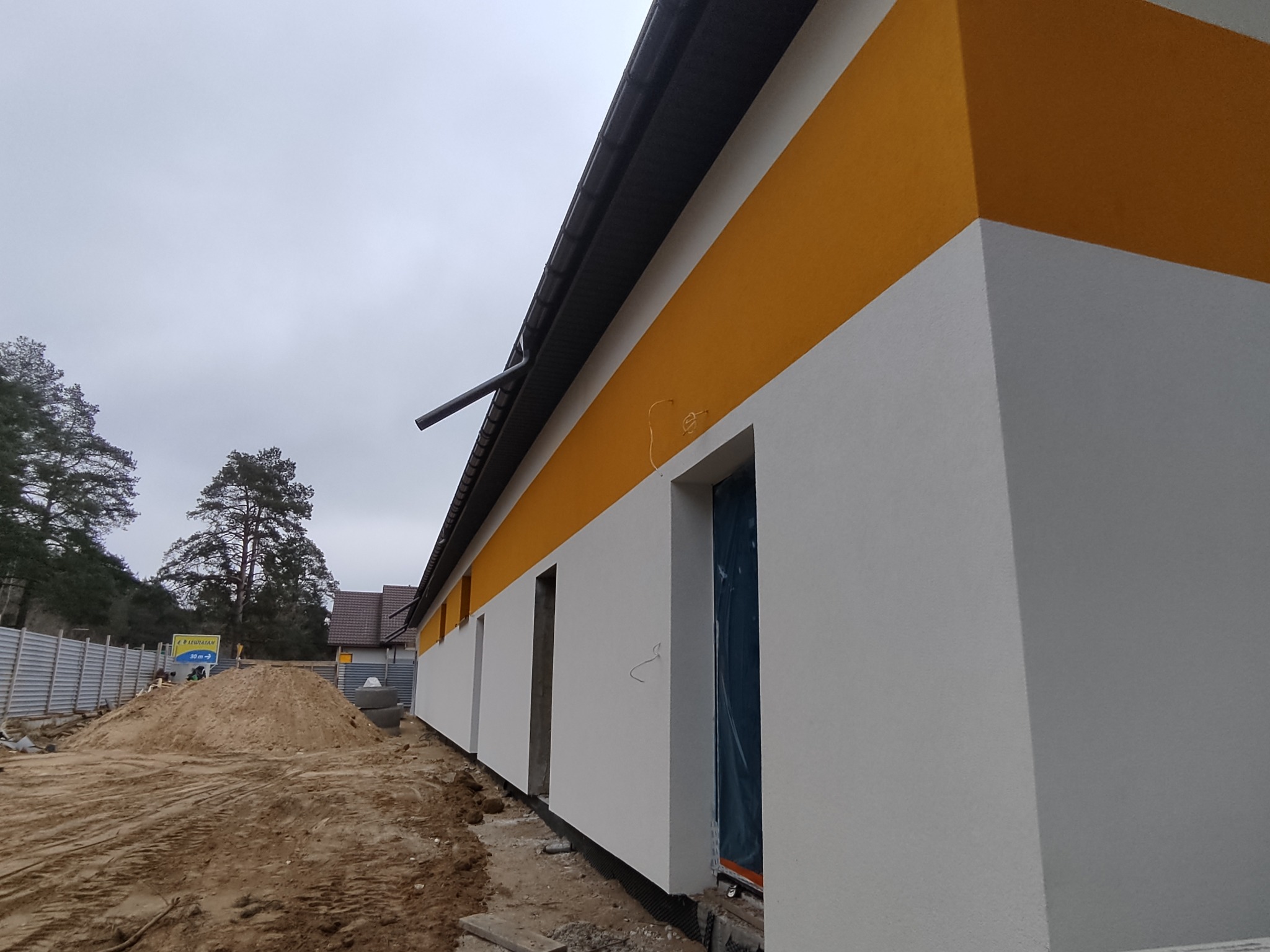 Świeżo otynkowana elewacja budynku z poziomym, żółtym pasem. Okna zabezpieczone folią. Na pierwszym planie hałda piachu, tło stanowią drzewa i szare niebo.