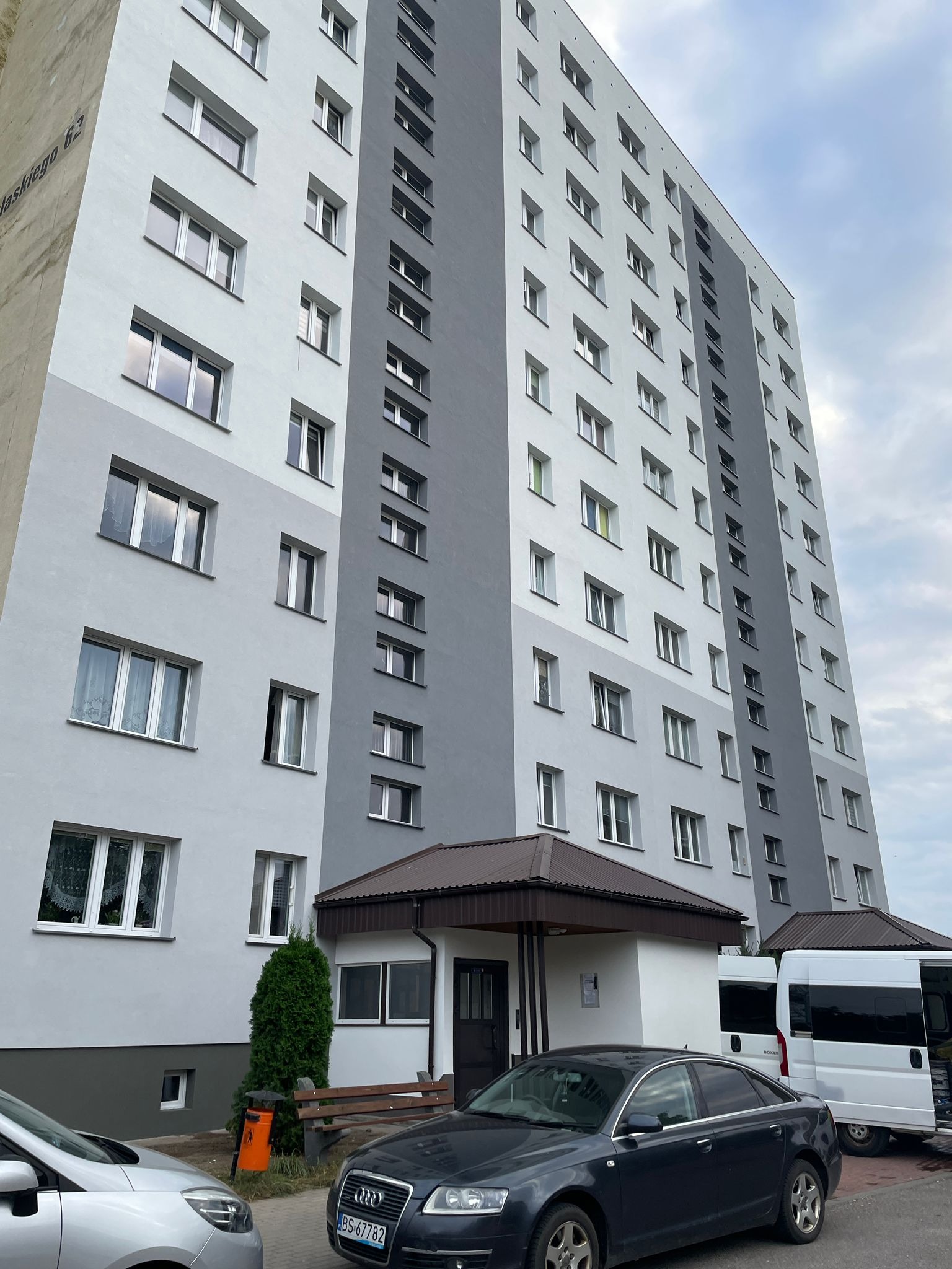 Wysoki budynek mieszkalny po renowacji elewacji, z widocznymi dwoma odcieniami szarości. Na pierwszym planie zaparkowane samochody i wejście do budynku.