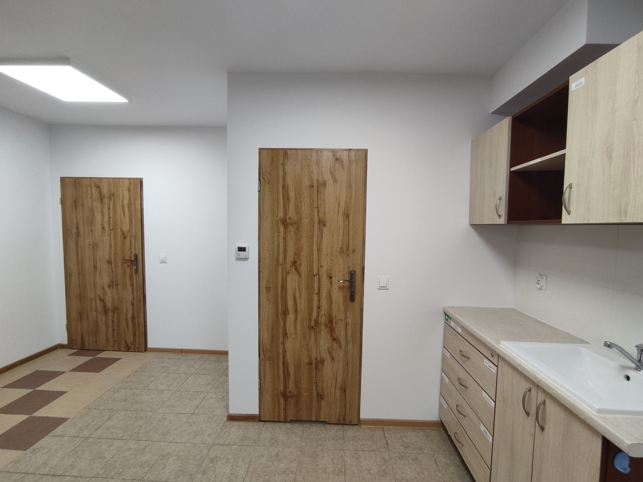 Wnętrze pomieszczenia po remoncie: jasne ściany, drewniane drzwi i szafki z umywalką. Podłoga wyłożona płytkami w odcieniach beżu i brązu. Oświetlenie sufitowe.