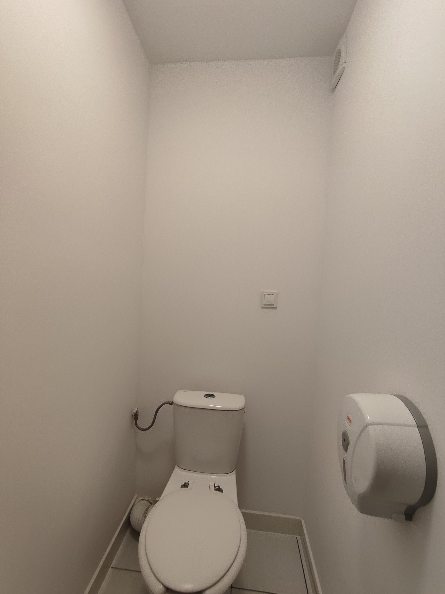 Biała toaleta w małej, odnowionej łazience z dozownikiem na ręczniki. Minimalistyczny design, jasne oświetlenie, czyste ściany. Widok z przodu.