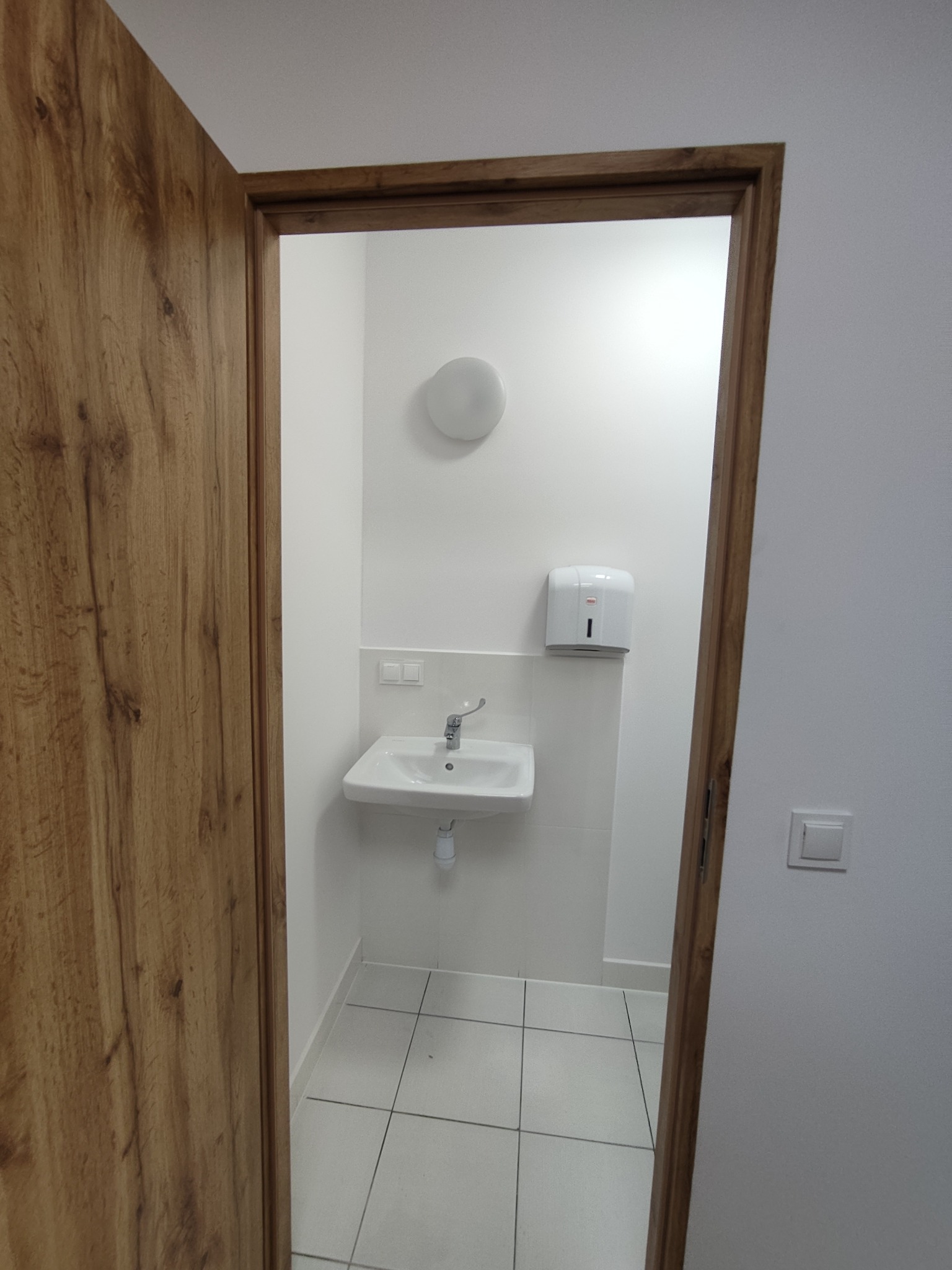 Biała łazienka po remoncie, widok z korytarza przez otwarte drewniane drzwi. Umywalka, dozownik mydła i białe płytki podłogowe. Minimalistyczny design.