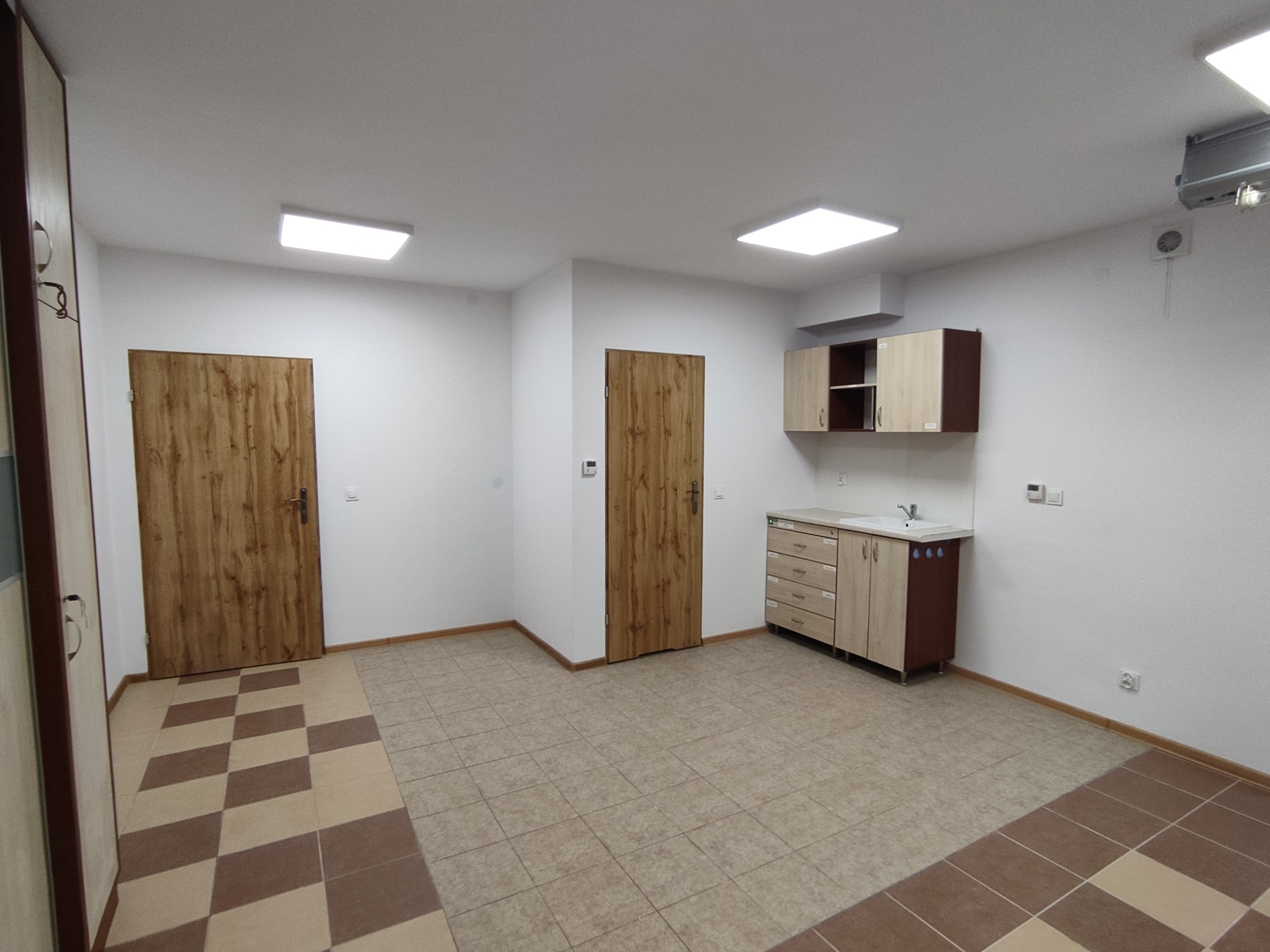 Wyremontowane wnętrze pomieszczenia z dwiema drewnianymi drzwiami, brązowo-beżową podłogą, szafkami i umywalką. Białe ściany i sufit z kwadratowymi lampami.