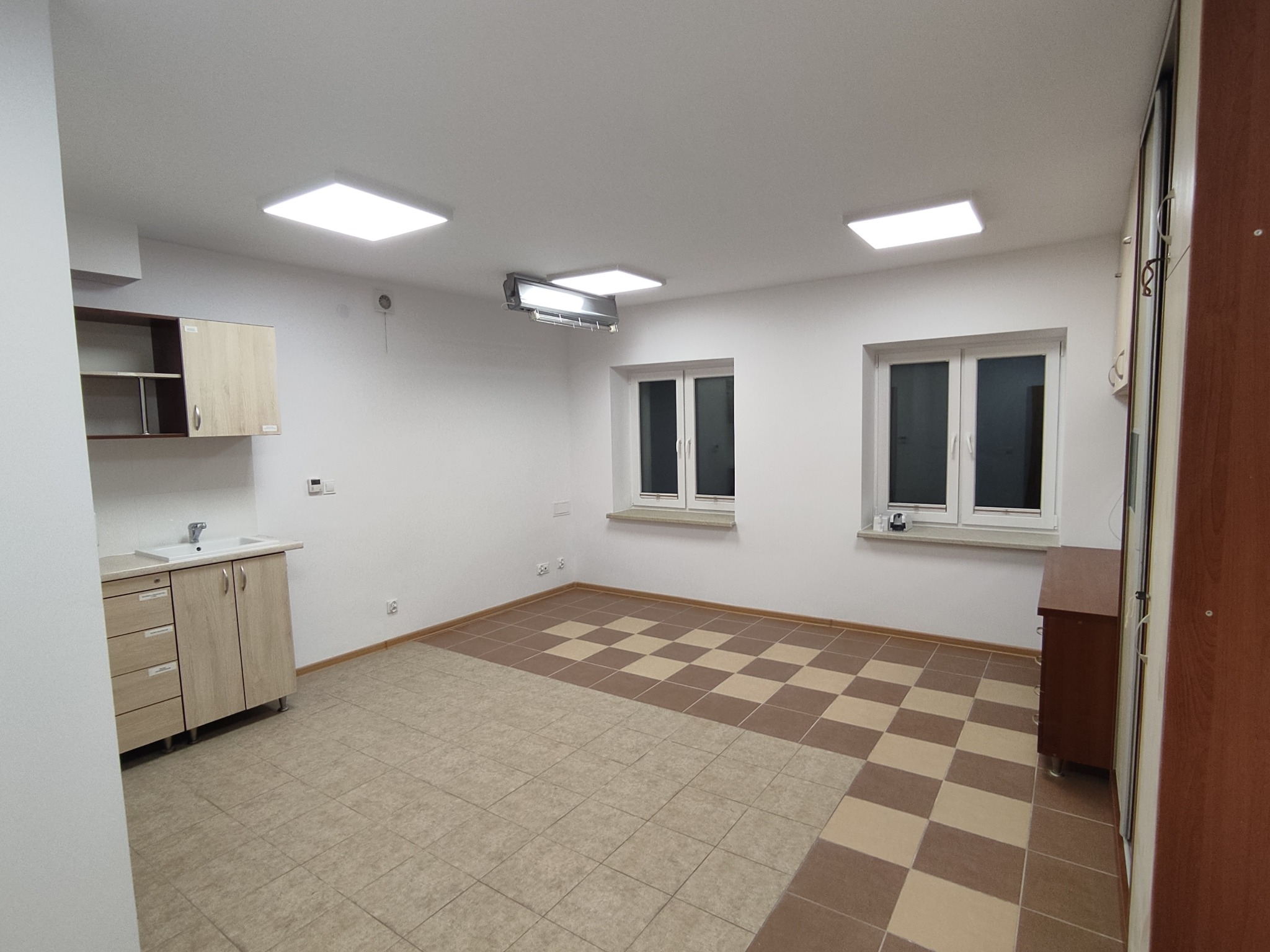 Wnętrze po remoncie: jasne ściany, kafelki w szachownicę, szafki z umywalką, okna z białymi ramami i nowoczesne oświetlenie sufitowe. Widok na odnowione pomieszczenie.