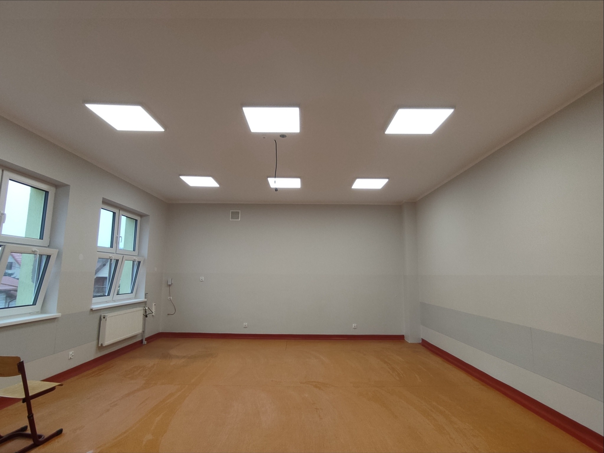Odnowiona sala edukacyjna z nowymi oknami, gładkimi ścianami w odcieniach szarości i równomiernie rozmieszczonymi, kwadratowymi lampami sufitowymi.