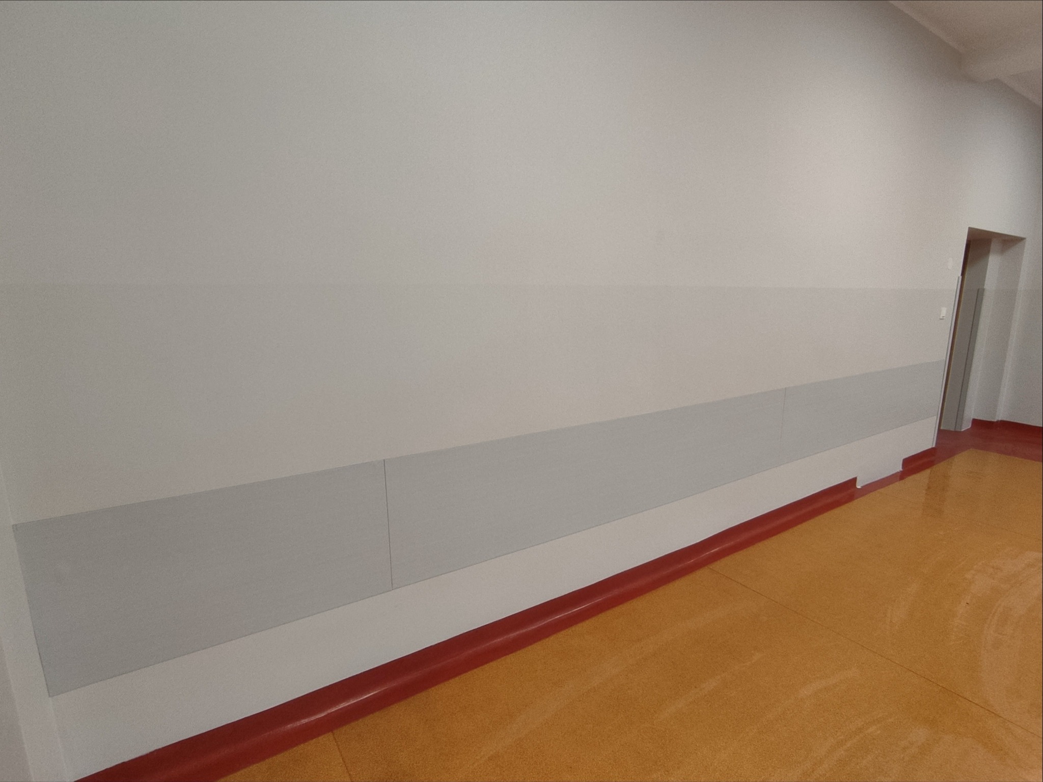 Wyremontowane wnętrze: jasne ściany z szarymi panelami ochronnymi na dole, czerwona listwa przypodłogowa i podłoga w odcieniach brązu. Widoczne drzwi w tle.