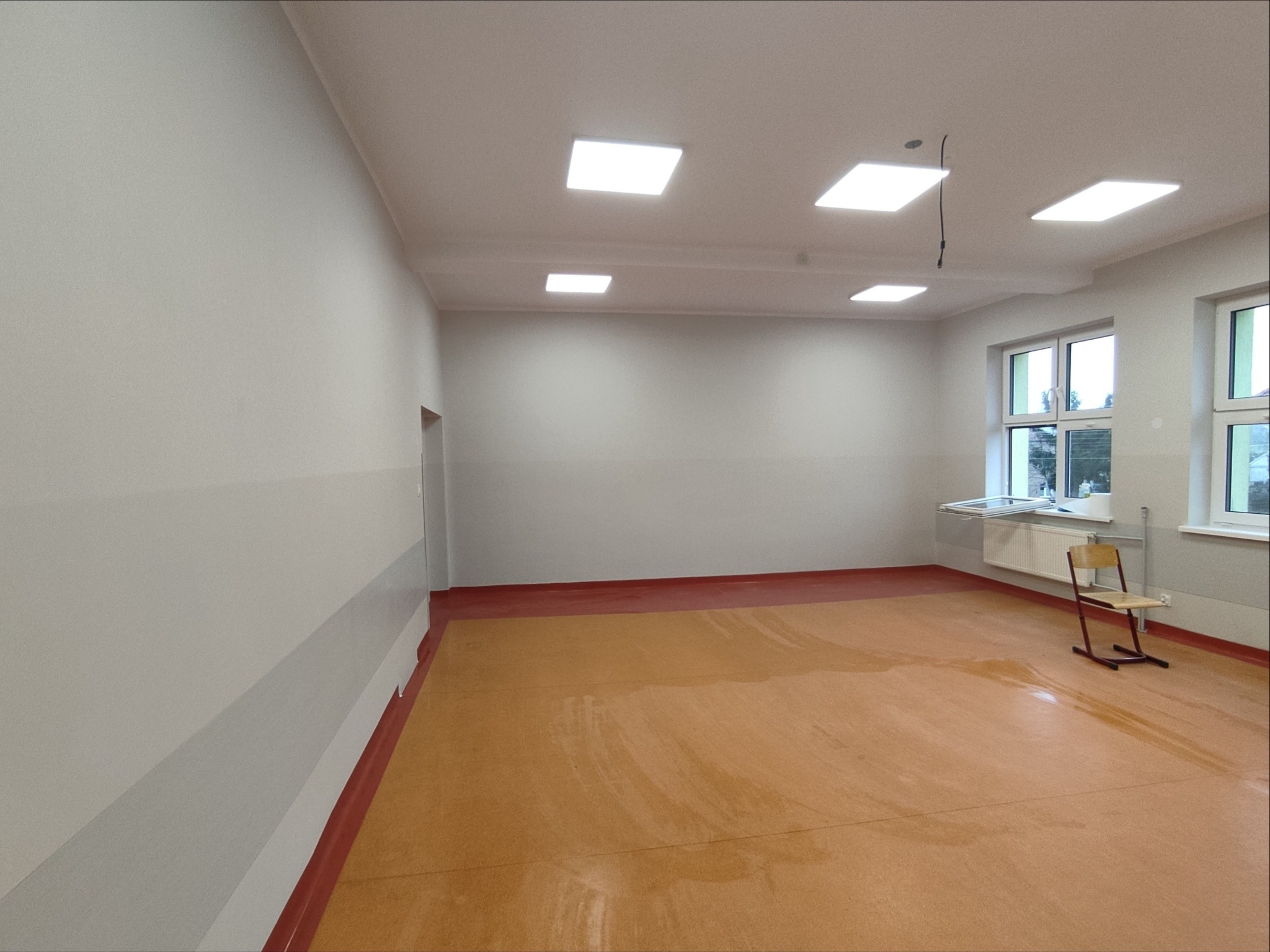 Wyremontowana sala edukacyjna z nowymi gładziami, pomalowanymi ścianami w odcieniach szarości i beżową podłogą. Widoczne okna i krzesło.