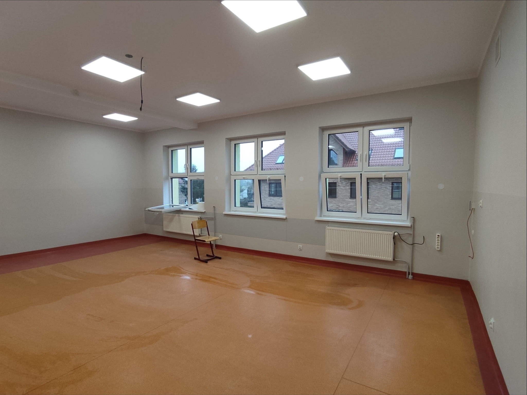 Wyremontowana sala edukacyjna z nowymi oknami, gładzią na ścianach, panelami LED i grzejnikami. Na podłodze widoczne ślady po remoncie. Krzesło na środku sali.