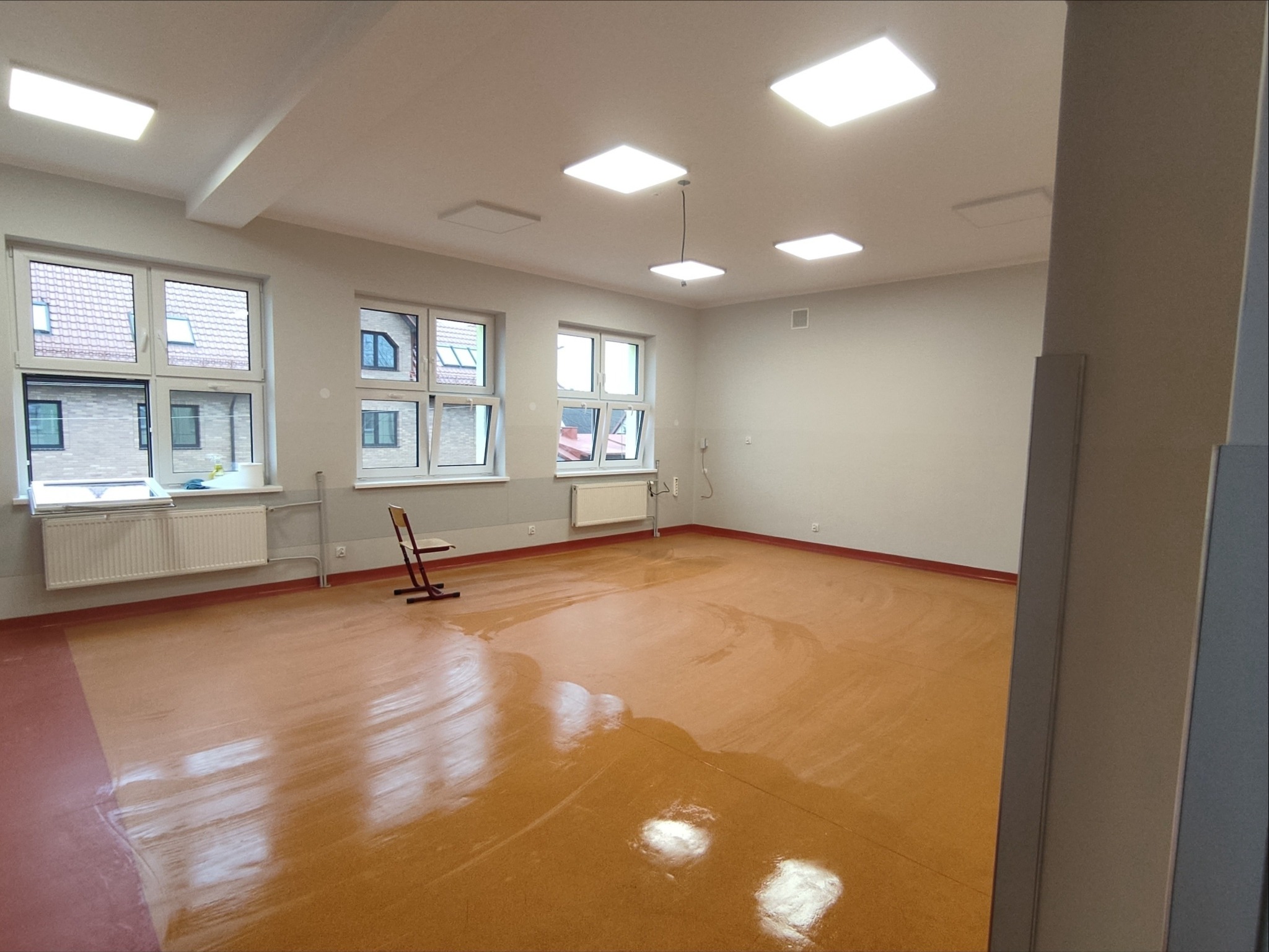 Wyremontowana sala edukacyjna z nowymi oknami, gładkimi ścianami i odnowioną podłogą. Widoczne grzejniki, oświetlenie sufitowe i krzesło. Świeżo po remoncie.