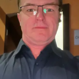 Portret mężczyzny w okularach i ciemnej koszuli na tle ściany w ciepłych odcieniach z elementami dekoracyjnymi.