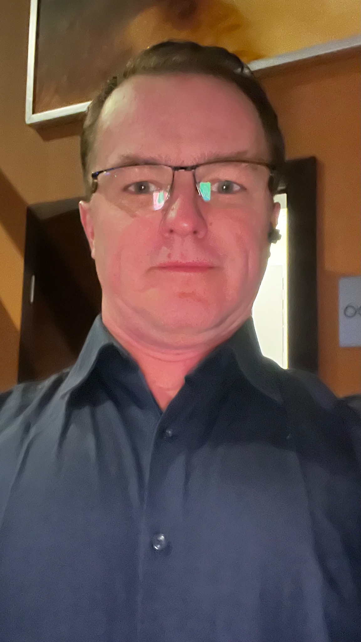 Portret mężczyzny w okularach i ciemnej koszuli na tle ściany w ciepłych odcieniach z elementami dekoracyjnymi.