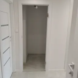 Wnętrze garderoby z szarymi płytkami podłogowymi i surowymi ścianami, widoczne przez białe drzwi z ościeżnicą.