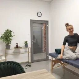 Kobieta siedzi na stole do masażu z wałkiem do ćwiczeń, w tle widoczny gabinet z roślinami i zegarem.