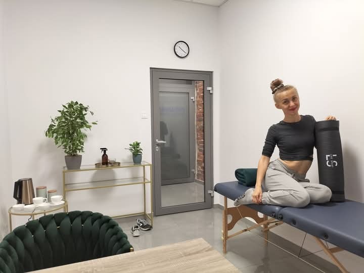 Kobieta siedzi na stole do masażu z wałkiem do ćwiczeń, w tle widoczny gabinet z roślinami i zegarem.