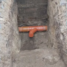 Pomarańczowa rura kanalizacyjna z trójnikiem ułożona w wykopie o nierównych ścianach, widoczne warstwy ziemi i fragmenty kostki brukowej na górze.