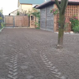 Przygotowany grunt pod trawnik z widocznymi śladami opon, ogrodzenie panelowe z podmurówką, drewniana altana z kwiatami w tle.