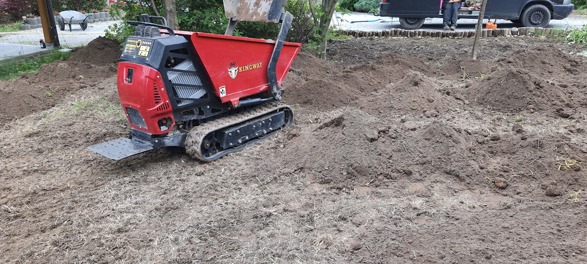 Czerwona mini wywrotka gąsienicowa marki Kingway na nierównym, przygotowywanym terenie z pryzmami ziemi, w tle widoczny fragment busa i wózek ogrodowy.