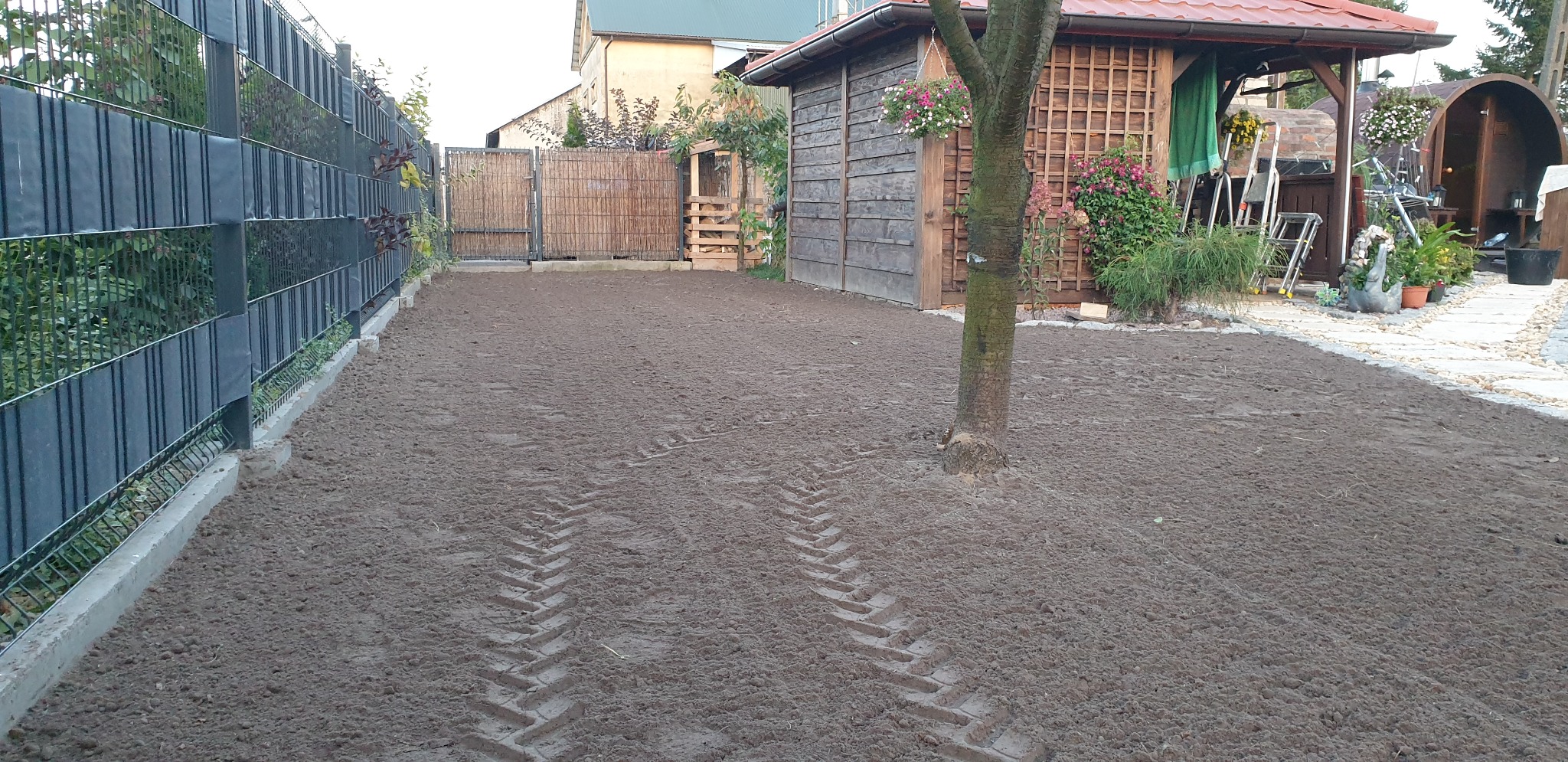 Przygotowany grunt pod trawnik z widocznymi śladami opon, ogrodzenie panelowe z podmurówką, drewniana altana z kwiatami w tle.
