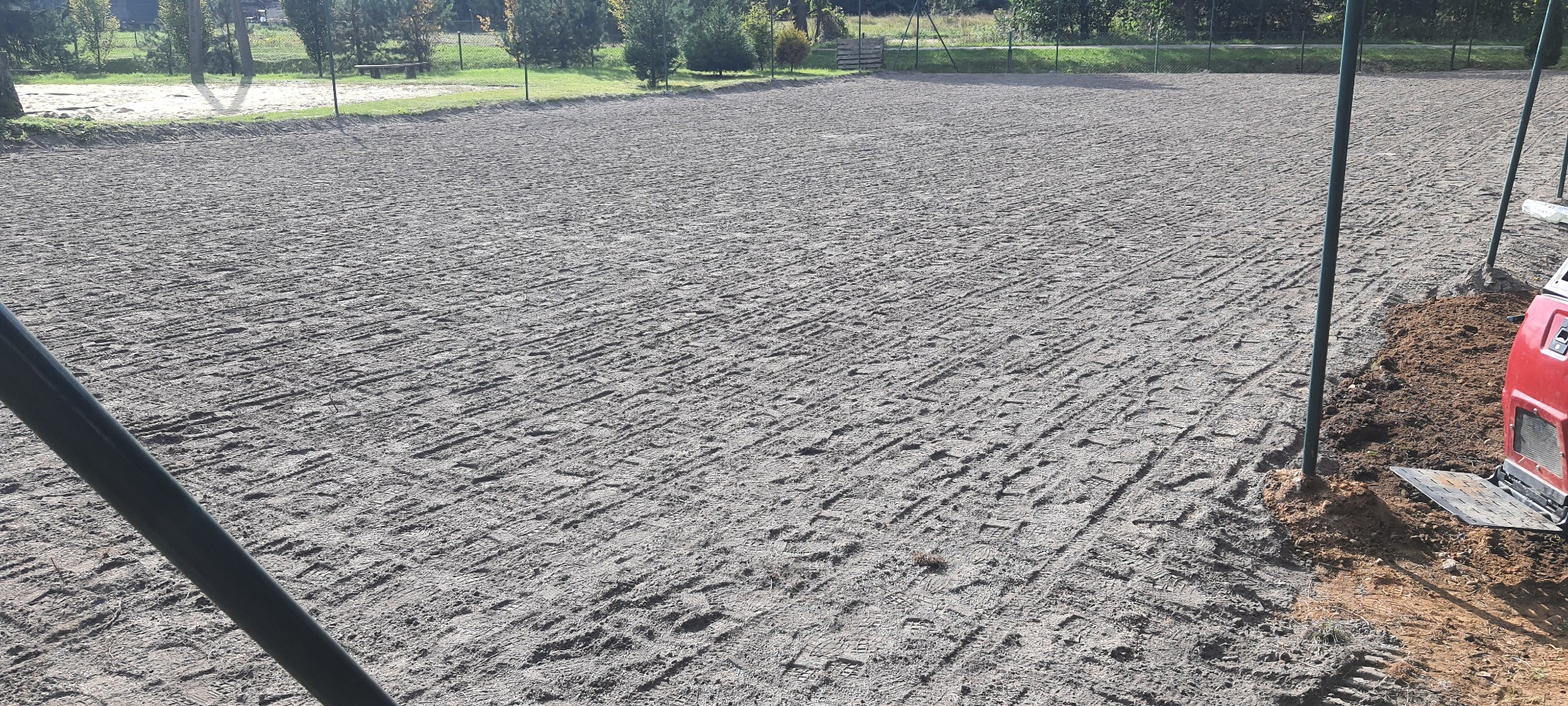 Równa, przygotowana powierzchnia ziemi z widocznymi śladami maszyn, otoczona zielenią i ogrodzeniem, z elementami maszyny budowlanej w prawym dolnym rogu.