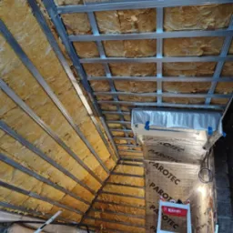 Skośny sufit poddasza w trakcie ocieplania, widoczne wełna mineralna między metalowymi profilami, folia paroizolacyjna z napisem PAROTEC, drabina i narzędzia.