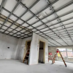 Sufit z widoczną metalową konstrukcją i częściowo zamontowanymi płytami izolacyjnymi, pomieszczenie w trakcie prac wykończeniowych, drabiny opierają się o ściany i framugi drzwiowe.