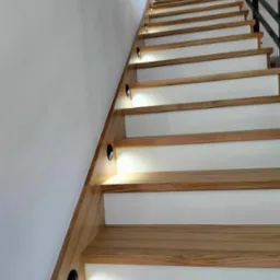Nowoczesne schody drewniane z białymi podstopnicami i oświetleniem punktowym w ścianie, widok z dołu.