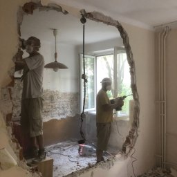 MADE2MEASURE SPÓŁKA Z OGRANICZONĄ ODPOWIEDZIALNOŚCIĄ - Dwóch pracowników w maskach ochronnych wykuwa otwór w ścianie, widoczne gruz i narzędzia, remont w trakcie.