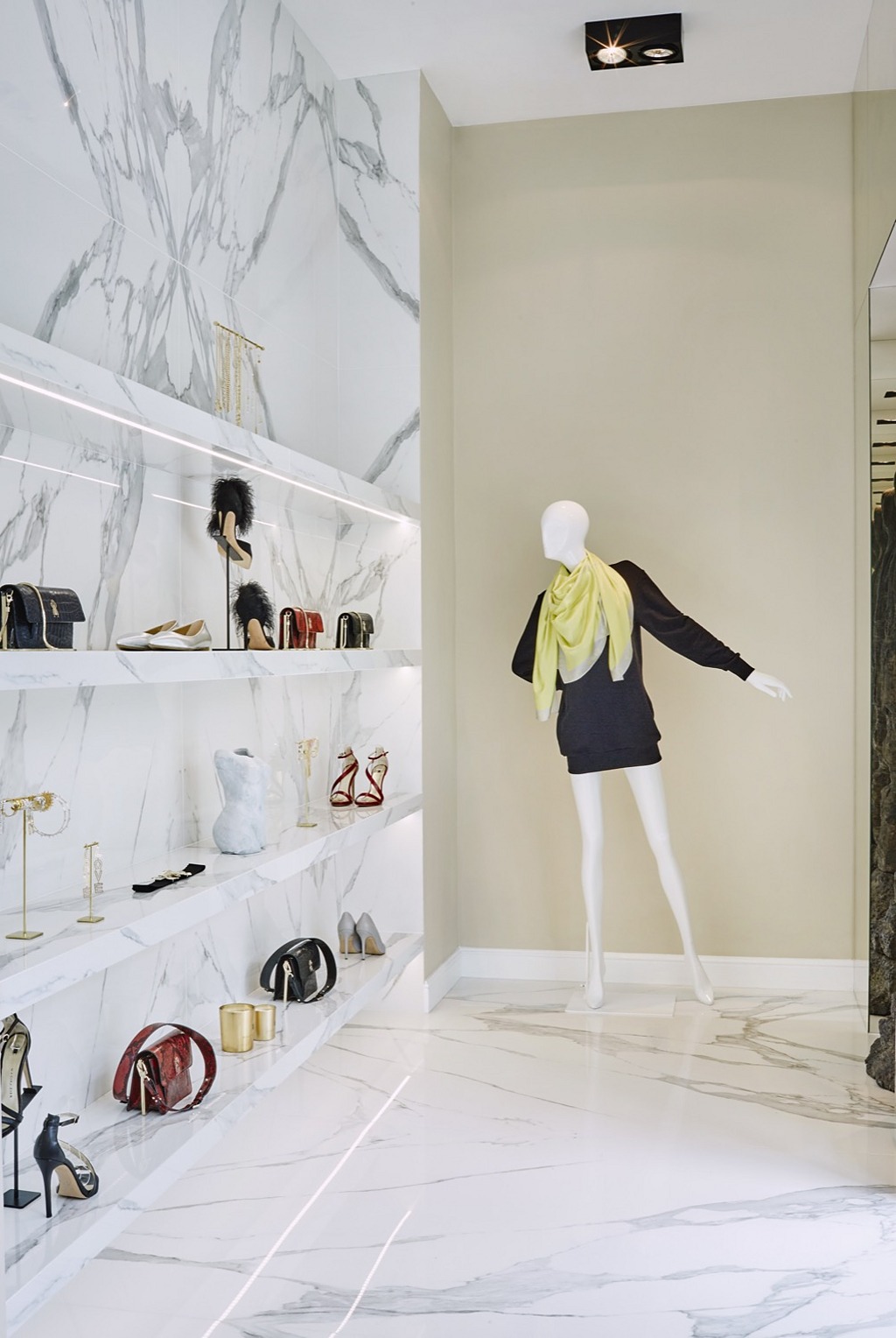 Wnętrze butiku z marmurowymi ścianami i podłogą, oświetlone półki prezentują torebki i buty, manekin z jasnożółtym szalem.