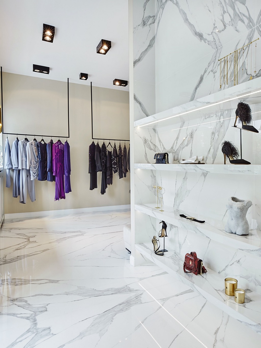 Wnętrze butiku z marmurową podłogą i ścianami, ubrania na wieszakach, półki z oświetleniem LED prezentujące torebki i obuwie, nowoczesne oświetlenie sufitowe.