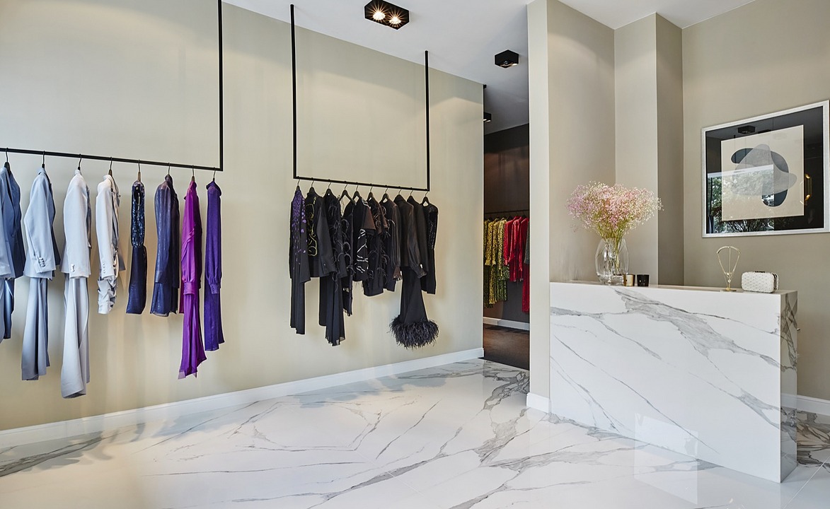 Wnętrze butiku odzieżowego z marmurową posadzką i ladą, wieszaki z ubraniami w odcieniach błękitu, fioletu i czerni, oświetlenie punktowe i dekoracje.