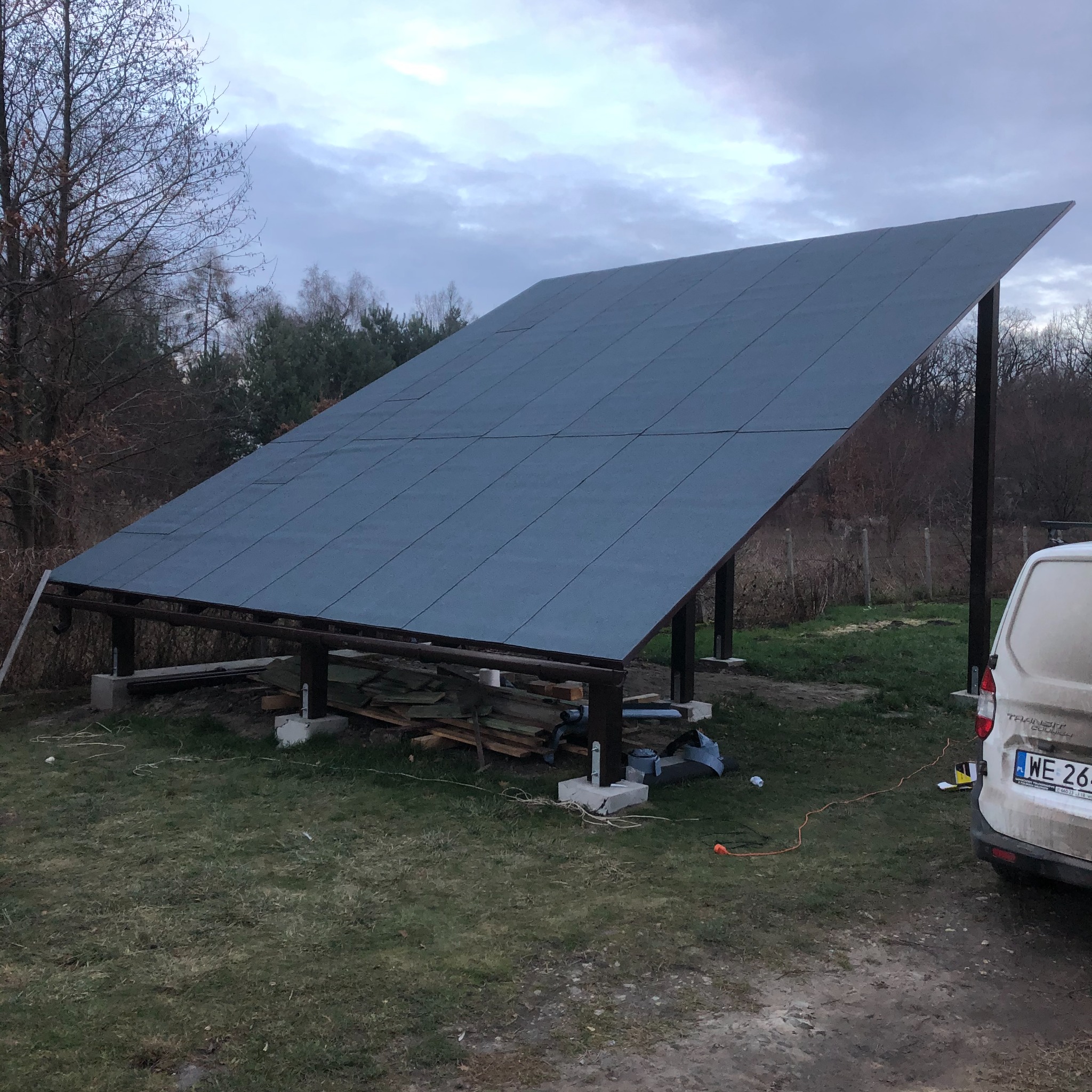 Konstrukcja z płyt na drewnianym szkielecie, prawdopodobnie zadaszenie tarasu, na zewnątrz, widoczny samochód dostawczy i przewody elektryczne.