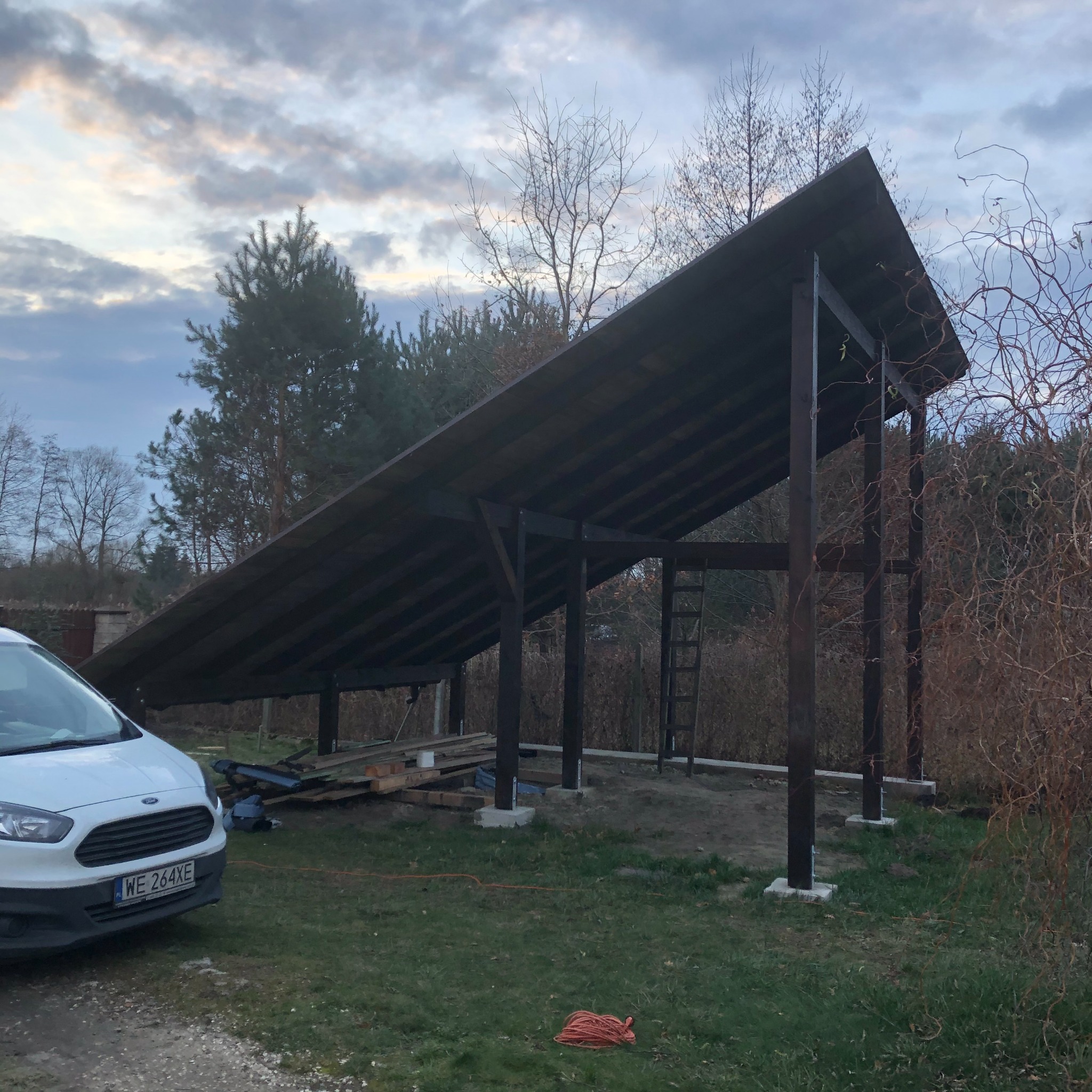 Konstrukcja drewnianej wiaty z dachem, wsparta na betonowych fundamentach, z widocznymi deskami i drabiną, obok zaparkowany biały samochód dostawczy Ford Transit z widoczną tablicą rejestracyjną