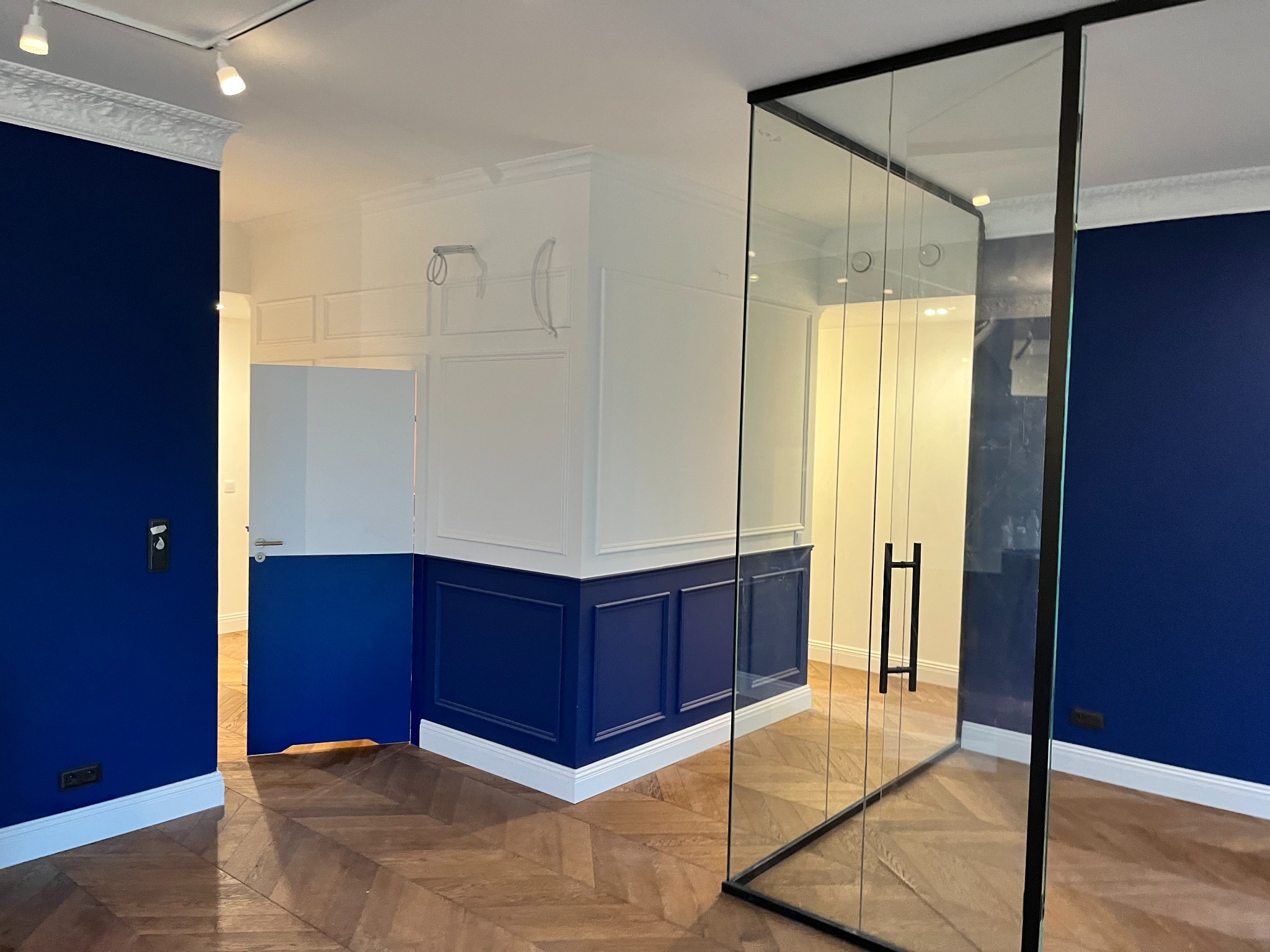 Wnętrze po renowacji z granatowymi ścianami, białymi listwami przypodłogowymi i sztukaterią, parkiet w jodełkę, przeszklona ścianka działowa z czarną ramą, otwarte drzwi pomalowane w połowie...